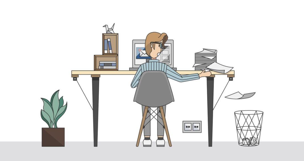 Employee at computer desk dropping papers into wire trash can.