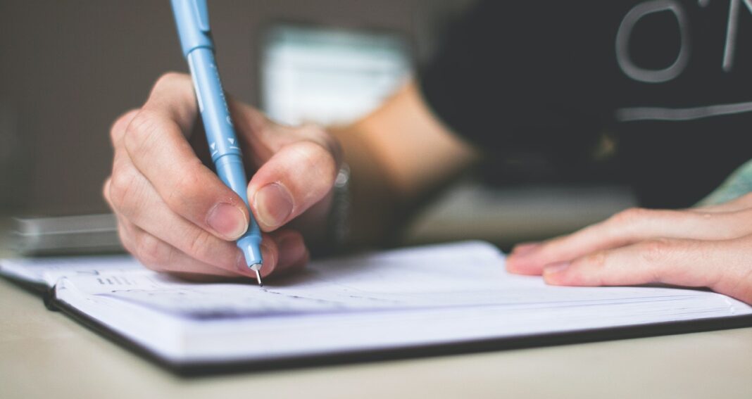 Person's arm and hand writing with a blue pen in a notebook.